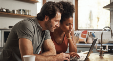 Two people seated at a kitchen counter reviewing content on a laptop, with notebooks and kitchen items visible nearby.